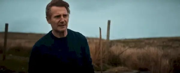 Liam Neeson filme šventųjų ir nusidėjėlių šalyje, jo personažas stovi priešais lauką ir atrodo susirūpinęs