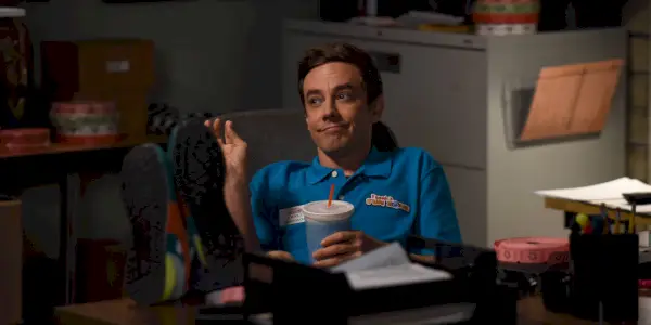 Jorma Taccone יושב מאחורי שולחן כשהוא מחייך בברוקלין תשע-תשע