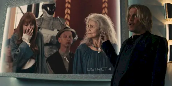 Haymitch ukazuje na obrazovku, která ukazuje, jak se Mags dobrovolně hlásí pro Annie v District 4 ve hře The Hunger Games Catching Fire