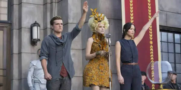 Peeta a Katniss pozdravujú tromi prstami, zatiaľ čo Effie hovorí na mikrofíne v Hunger Games Catching Fire