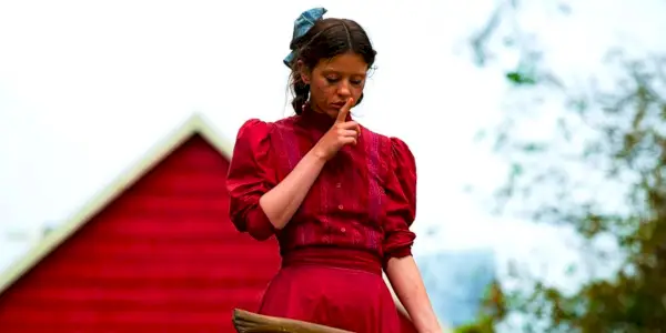Mia Goth con un vestido rojo haciendo callar a alguien mientras sostiene un hacha en Pearl