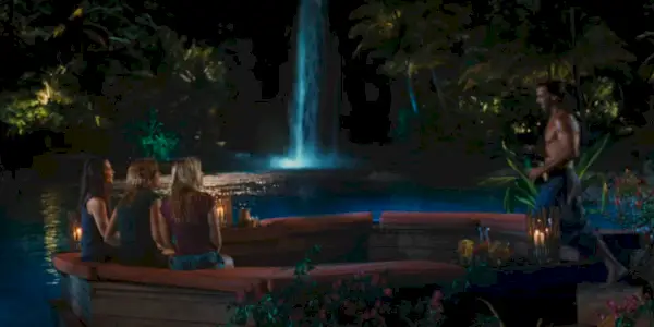 Três meninas sentam-se em frente a uma cachoeira com um homem caminhando em sua direção.