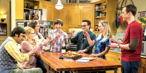 Johnny Galecki als Leonard, Melissa Rauch als Bernadette, Jim Parsons als Sheldon, Kunal Nayyar als Raj, Kaley Cuoco als Penny, Mayim Bialik als Amy und Simon Helberg als Howard stoßen in „The Big Bang Theory“ alle mit Champagner an einem Tisch an