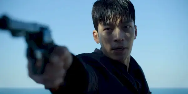 Jun-ho aponta sua arma contra o céu azul e o oceano ao longe no programa Squid Game da Netflix.
