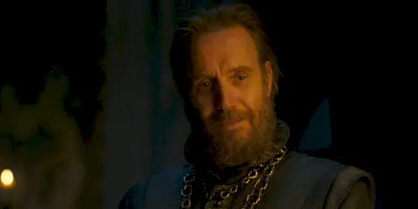 Otto Hightower sorrindo suavemente em frente à lareira em House of the Dragon.