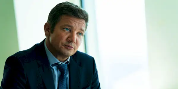 Jeremy Renner como Mike McClusky sentado a la mesa y vistiendo un traje en el episodio 6 de la temporada 2 del alcalde de Kingstown.