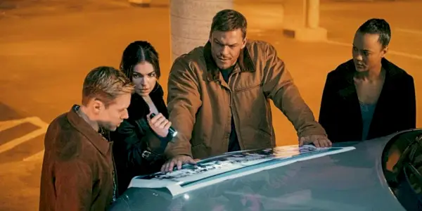 Reacher rodeado por el elenco de la nueva temporada 2 mirando algunos documentos en el capó de un auto.