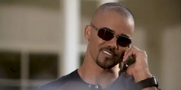 תמונה של דרק מורגן מדבר בטלפון ב-Criminal Minds, מרכיב משקפי שמש ומחייך.