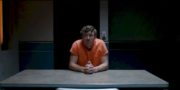 Zach Gilford sentado com um macacão de prisão em uma sala vazia em Criminal Minds Evolution