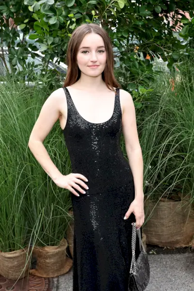 Kaitlyn Dever, eine junge Frau, blickt lächelnd in die Kamera und posiert mit der Hand auf der rechten Hüfte. Sie hat dunkelbraunes Haar, das glatt nach unten getragen wird, und trägt ein langes schwarzes Paillettenkleid