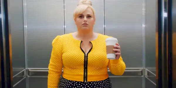 Rebel Wilson dans un haut jaune vif et tenant une tasse de café dans l'ascenseur dans Isnt It Romantic