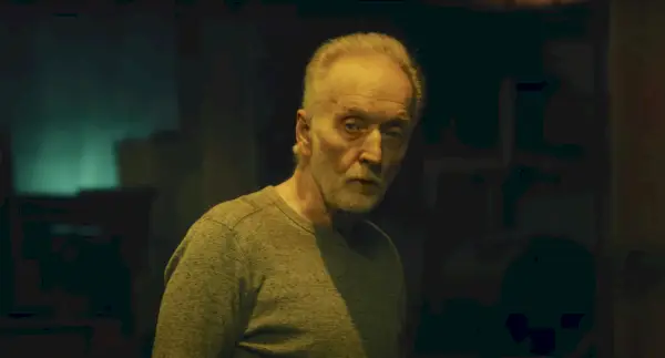 Tobin Bell como rompecabezas en Saw X, un hombre mayor con cabello y barba grises se encuentra en una habitación oscura mirando a la cámara, lleva una blusa gris de manga larga.