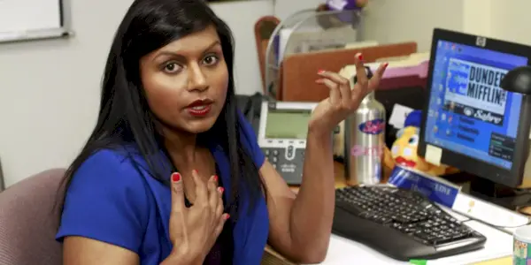 Kelly Kapoor med hånden på brystet ved skrivebordet på kontoret