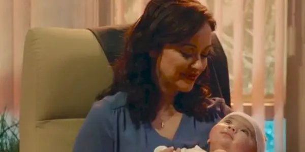 Blue Bloods Baez hält ihr Baby. Sie sitzt auf einem grünen Stuhl, trägt ein blaues Hemd und blickt auf den Säugling herab. Hinter ihr sind offene Jalousien.